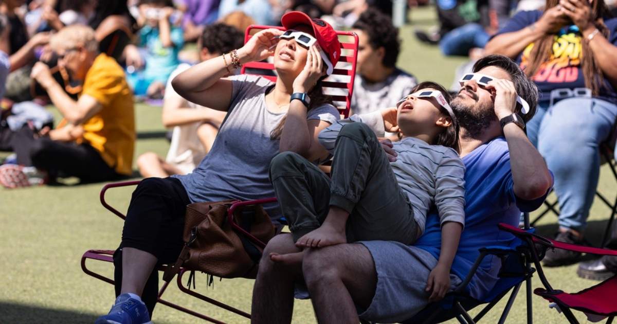 Familie kijkt met zonnebrillen voor zonsverduistering omhoog naar de lucht, zittend op campingstoelen tussen een menigte mensen buiten