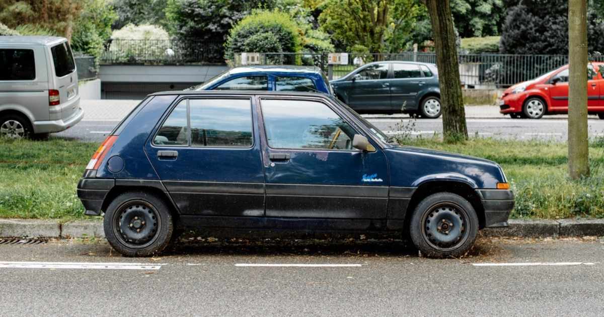 Donkerblauwe oude hatchback geparkeerd langs de stoeprand, met op de achtergrond andere auto's en groen