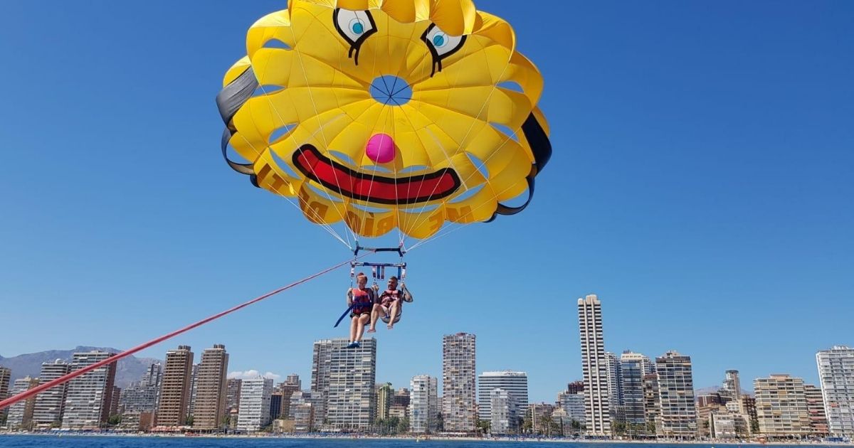 Twee personen parasailen boven Benidorm met een gele clownsparachute, met de skyline van de stad op de achtergrond