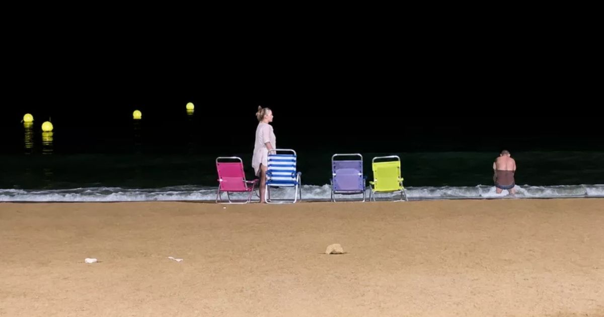 Twee mensen op een verlaten strand 's nachts, met vier kleurrijke stoeltjes aan de waterlijn en gele boeien in zee