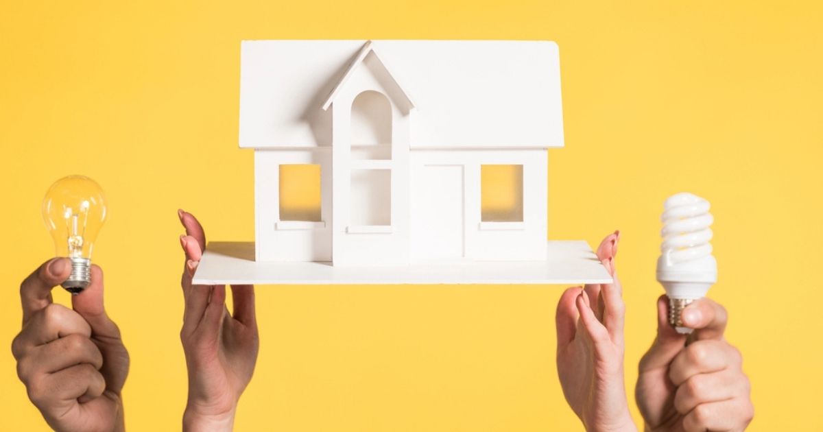 Handen houden een wit miniatuurhuis omhoog, geflankeerd door een gloeilamp en een energiezuinige spiraallamp