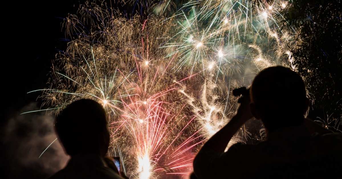 Twee mensen kijken naar een kleurrijk vuurwerk aan de nachtelijke hemel, één fotografeert het spektakel